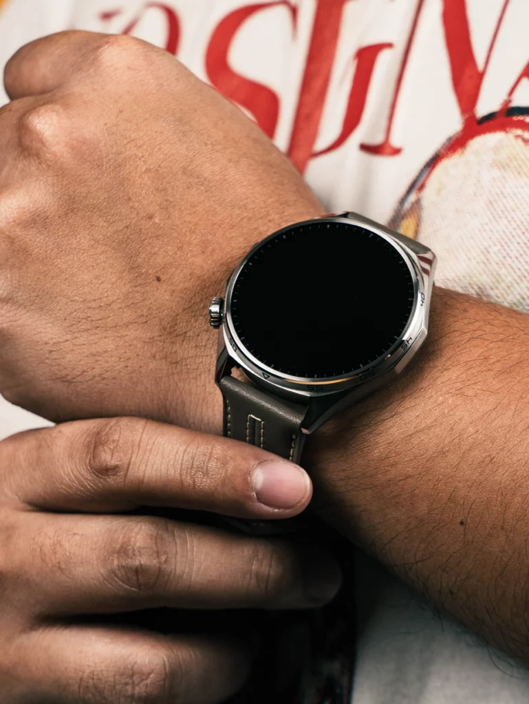 Close-up on a person's wrist wearing the Huawei Watch GT 6 Pro with a dark leather strap. The watch face is off (black), and the person's hands are visible near the watch.
