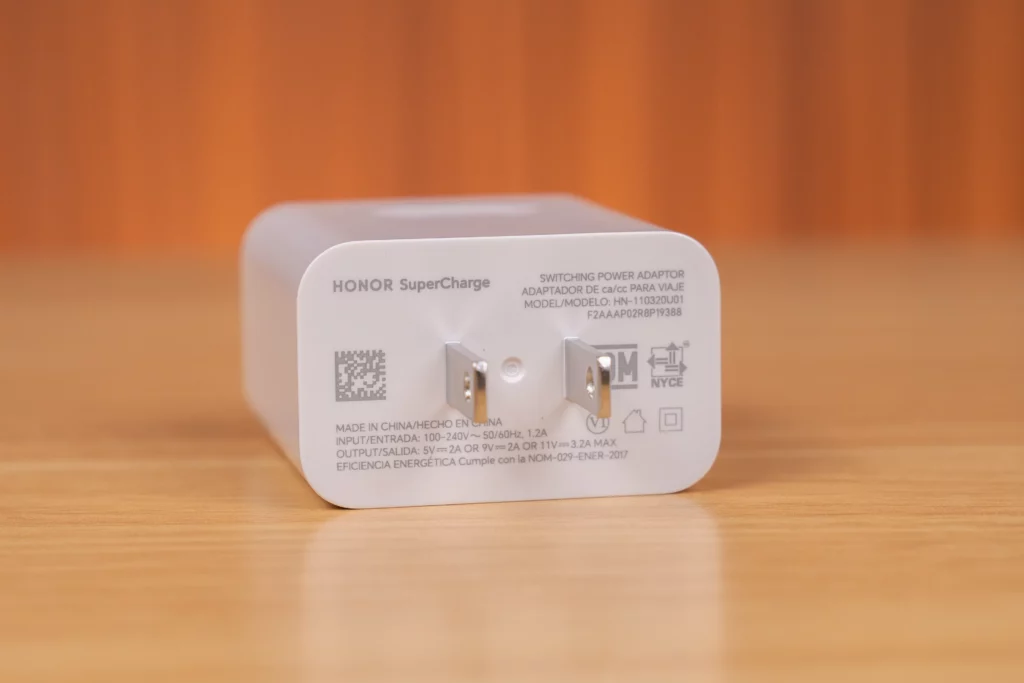 Close-up of the white HONOR SuperCharge power adapter. The label shows input voltage (100-240V) and output ratings, including 5V=2A, 9V=2A, or 11V=3.2A MAX. It has a two-pronged US-style plug.