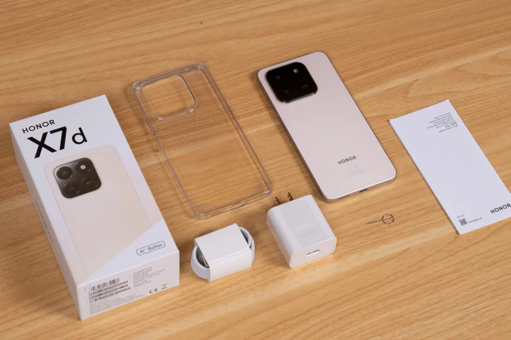 Unboxing contents of the HONOR X7d. Laid out next to the box are the light-colored phone, a clear silicone case, a white charging adapter, a white USB cable, a SIM ejector tool, and documentation.