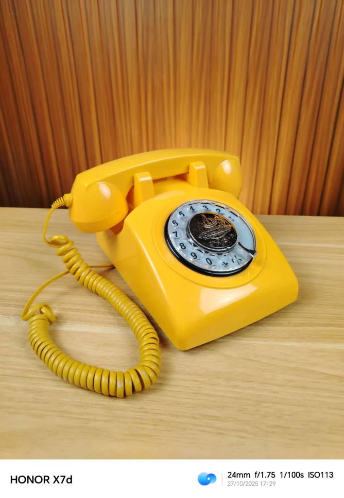 A photograph taken by the HONOR X7d (indicated by the watermark at the bottom). The image shows a bright yellow retro rotary telephone with a coiled cord, sitting on a light wooden table with a vertically-striped background. The camera's settings are watermarked as 24mm f/1.75 1/100s ISO113.