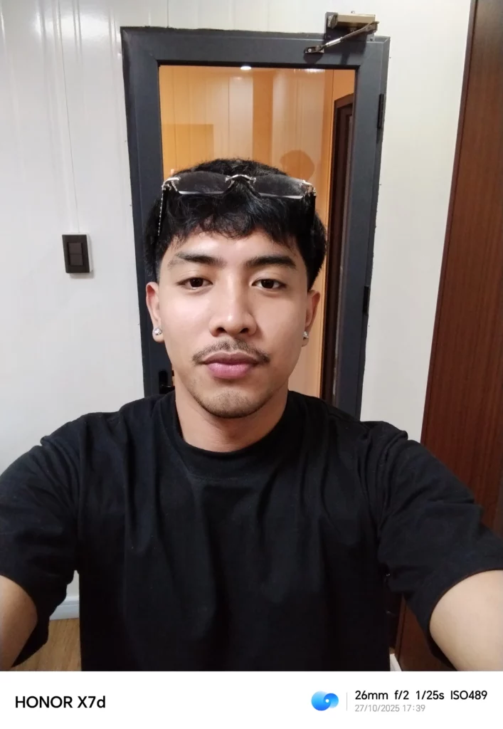 A selfie-style portrait of a man with a mustache and a black shirt, looking directly at the camera. He is wearing glasses on top of his head. The background shows a white wall and a doorway with a dark frame.