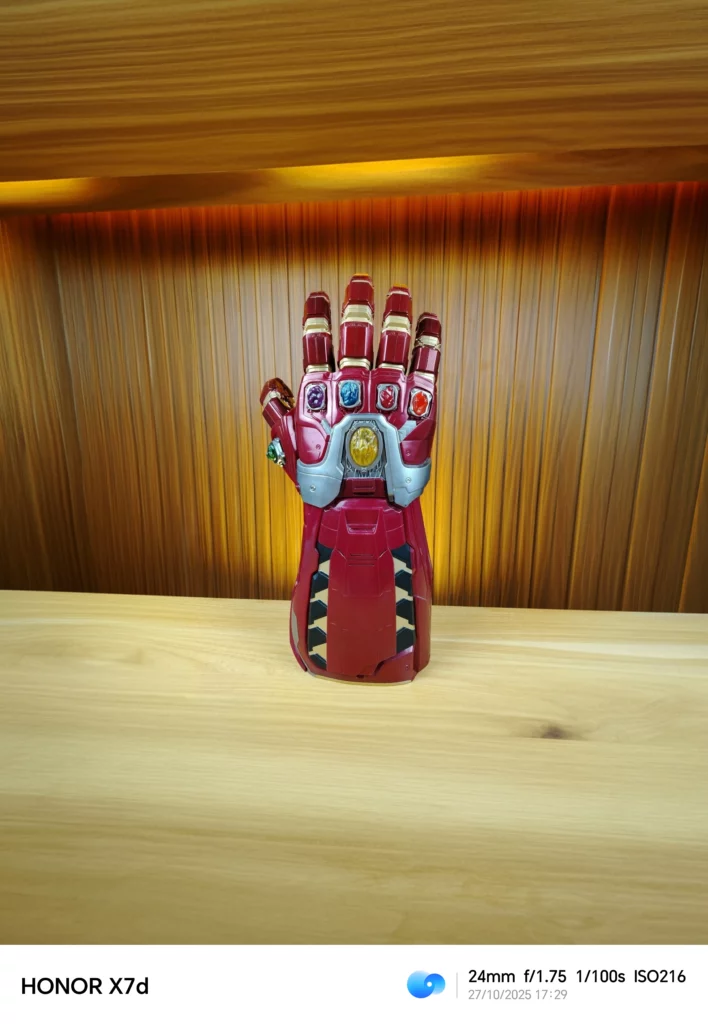 A slightly wider shot of a red and gold Iron Man-themed Infinity Gauntlet toy, showing more of the wooden table and the wooden paneled background above it, which has warm, upward-shining ambient lighting.