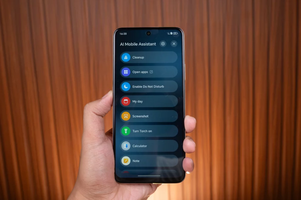 A smartphone screen is displayed, showing the "AI Mobile Assistant" menu with a list of quick actions and tools, including Cleanup, Open apps, Enable Do Not Disturb, My day, Screenshot, Turn Torch on, Calculator, and Note. The phone is held in a hand against a vertically-paneled wooden background.