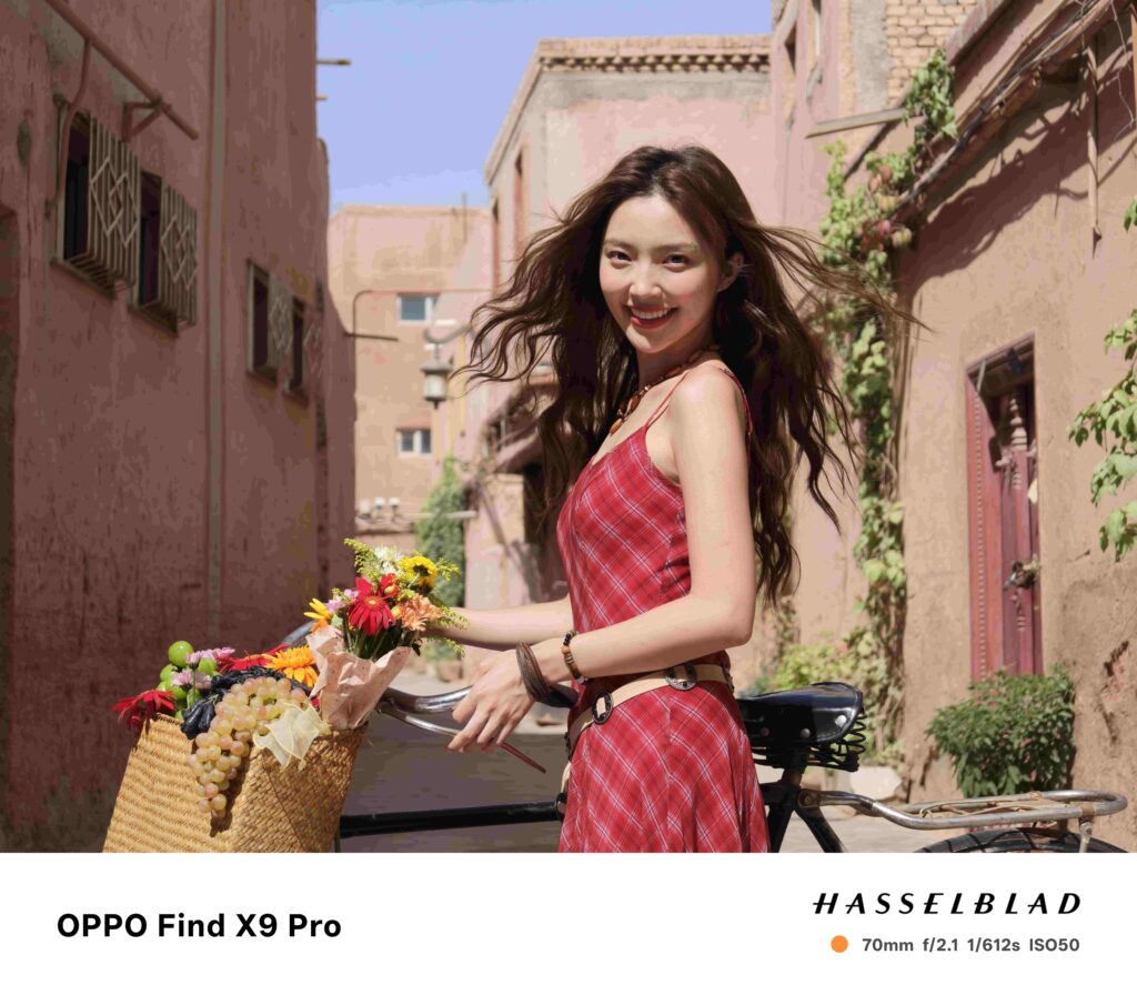 A bright sample photograph taken with the OPPO Find X9 Pro. The image features a smiling woman in a red plaid dress standing next to a bicycle with a basket full of flowers and grapes. She is positioned on a narrow street with reddish-orange buildings in the background. The photo is labeled OPPO Find X9 Pro and includes HASSELBLAD branding with camera metadata: 70mm f/2.1 1/612s ISO50.