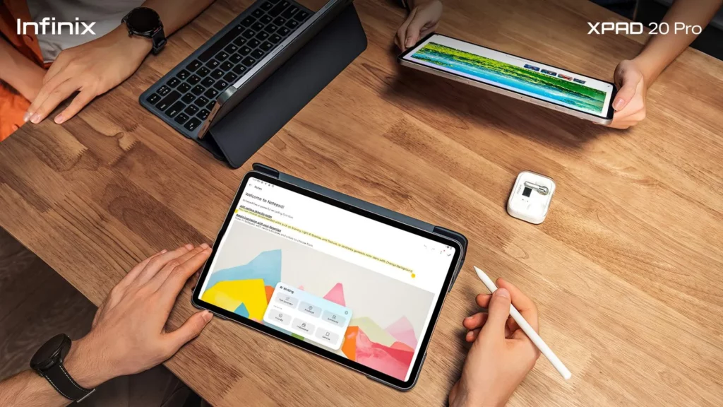 A close-up, overhead shot of three people collaborating around a wooden table. Two Infinix XPAD 20 Pro tablets are visible: one is horizontal with a keyboard case, showing a colorful document or app, while the other is held by a person and shows a spreadsheet or web page. A stylus is held over the main tablet.