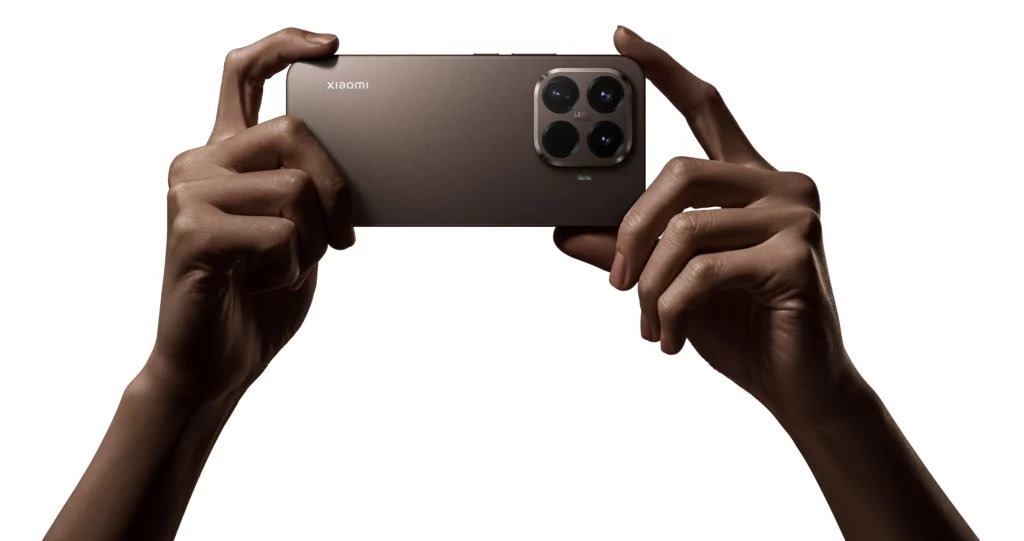 A close-up of a person holding the back of a brown Xiaomi 15T Pro smartphone horizontally with both hands. The large camera module with four lenses and the Leica branding is visible. The background is white.