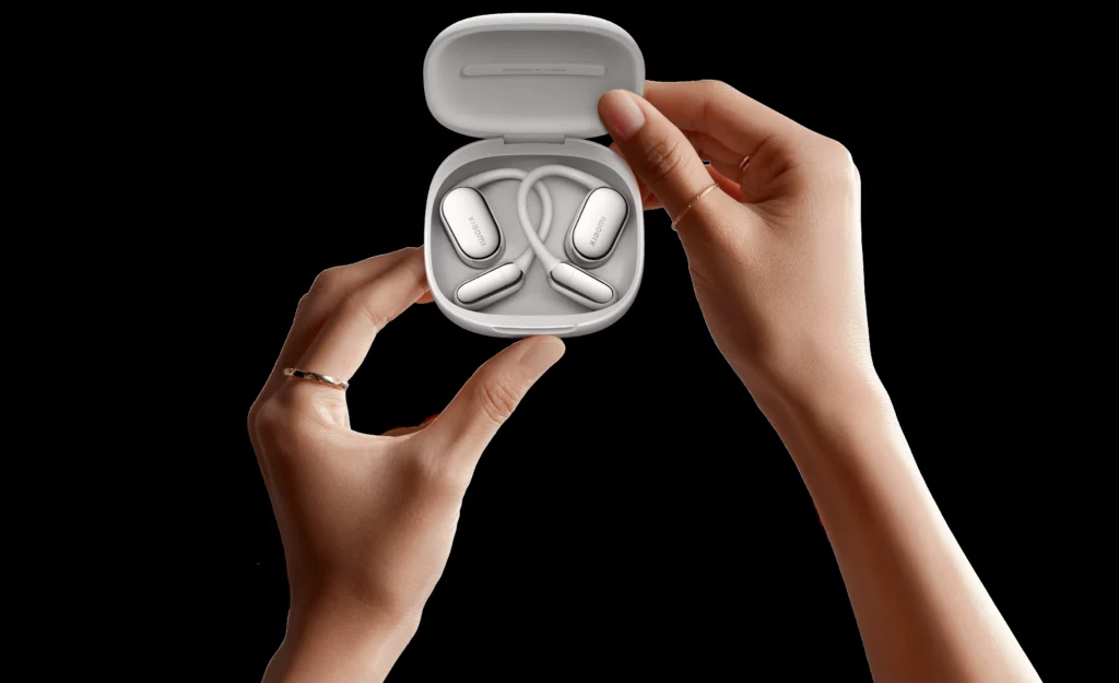 A close-up of hands holding the open charging case for the silver Xiaomi Openwear Stereo Pro earbuds. The earbuds feature an open-ear design with an ear hook/band connecting the two buds. The background is black.