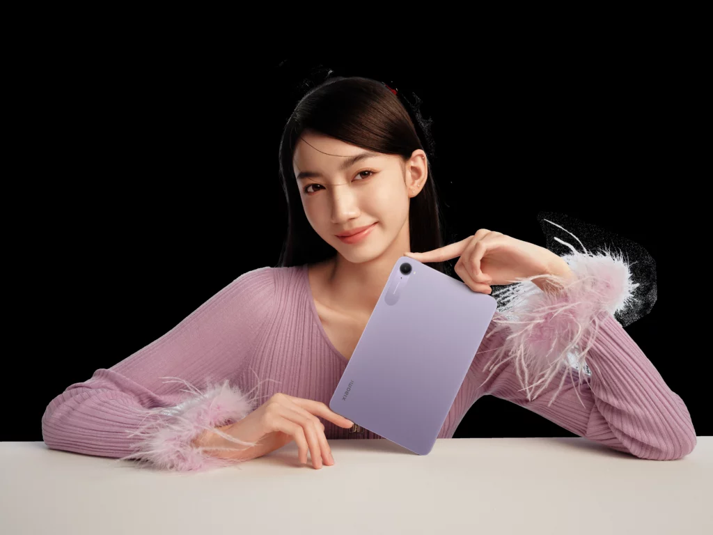 A portrait shot of a smiling woman holding the Xiaomi Pad Mini in a light purple color. She is wearing a soft pink sweater with feathery cuffs, and the background is black.