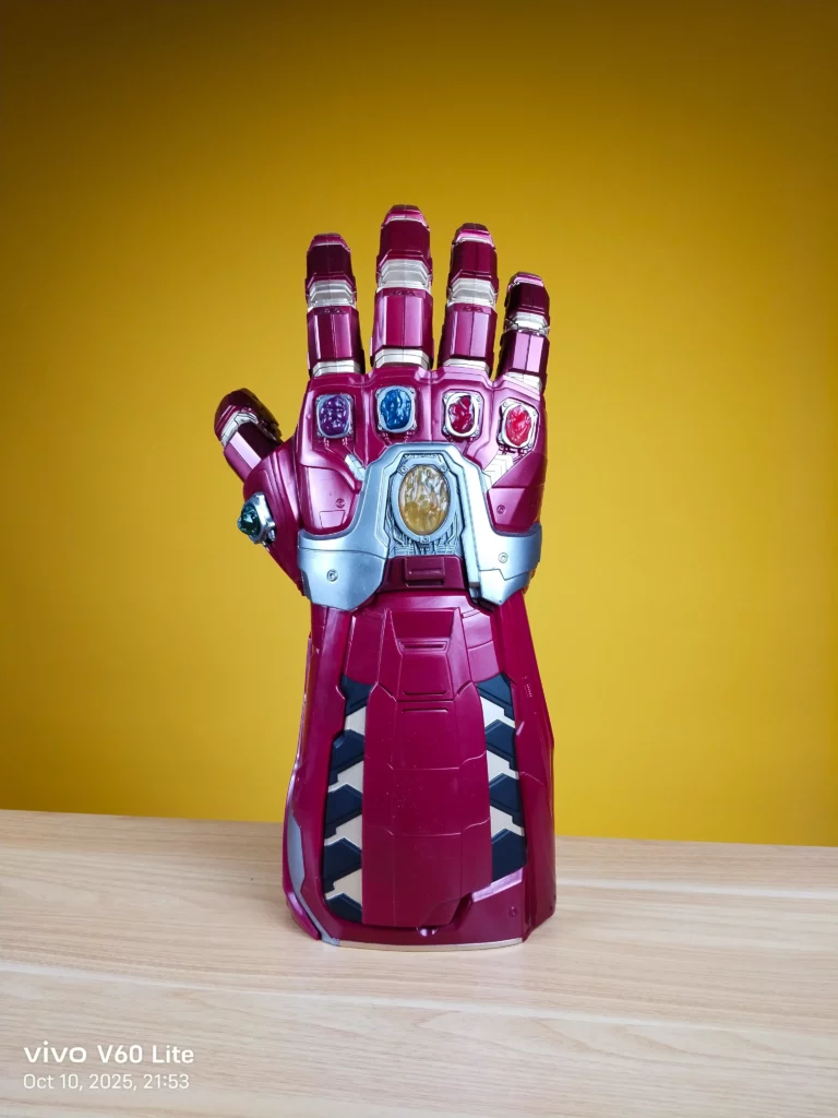 A photo, with a camera watermark indicating it was taken on the vivo V60 Lite, showing a large, plastic, crimson-colored Iron Man Infinity Gauntlet replica toy standing upright against a plain yellow background.