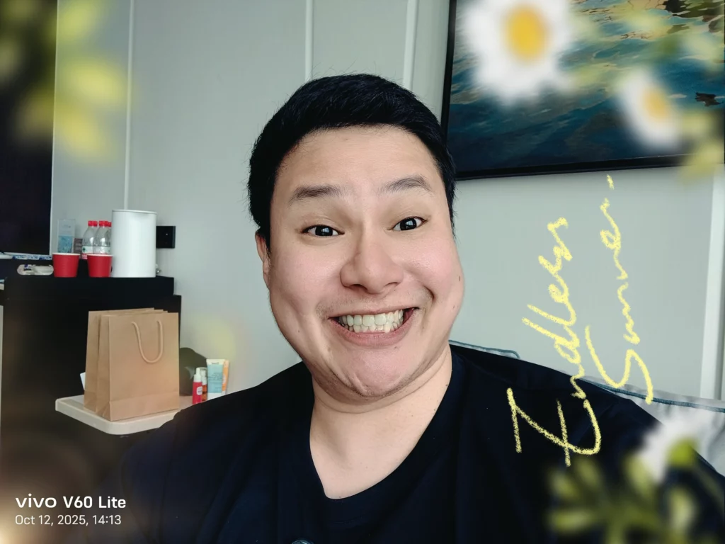 A selfie photo, with a camera watermark indicating it was taken on the vivo V60 Lite, showing the same man from the previous photo with a wide smile, but this image has been edited with a filter that includes light flares and yellow digital scribbles (possible signature/text) overlaid on the image.
