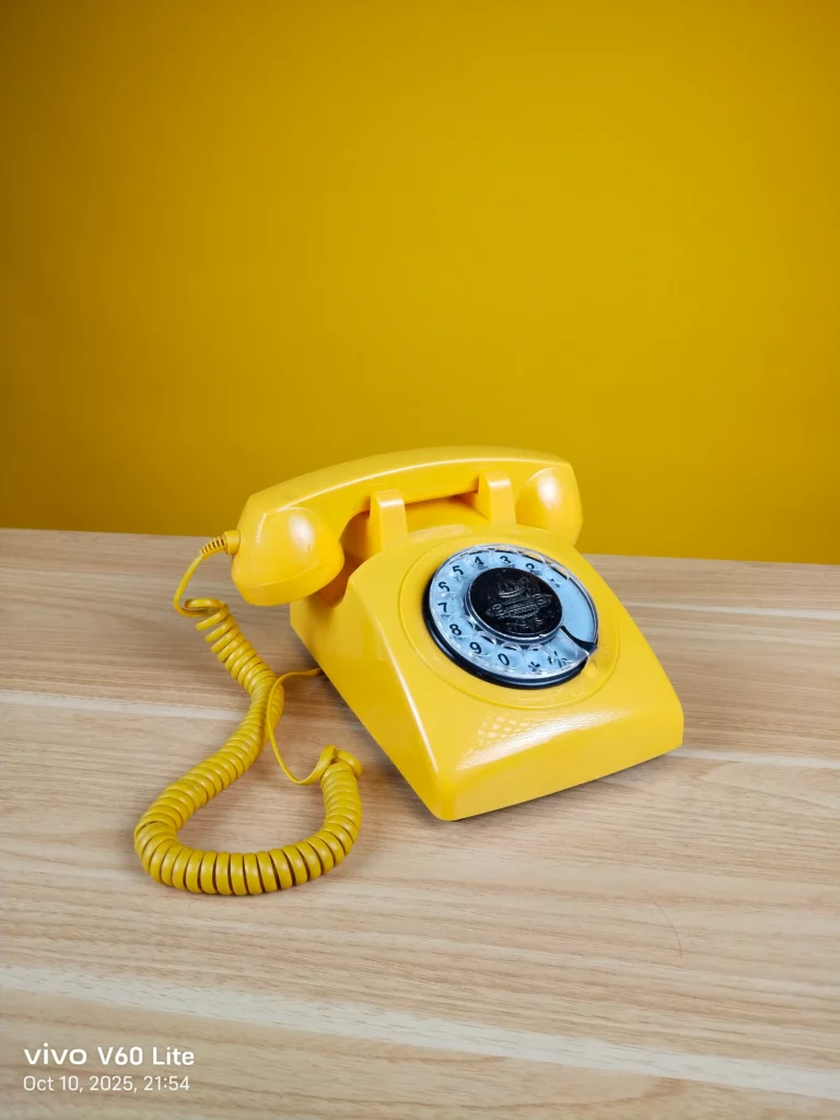 A photo, with a camera watermark indicating it was taken on the vivo V60 Lite, showing a classic, brightly yellow rotary telephone resting on a light wooden surface against a bold yellow background. The coiled cord is visible.
