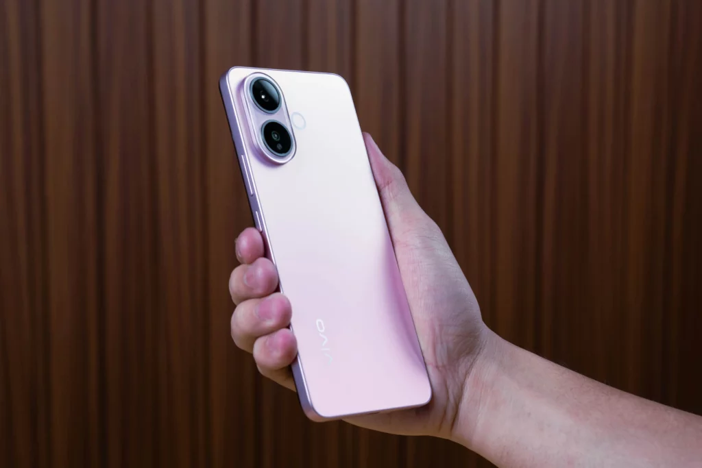 A person's hand holds the light purple vivo V60 Lite phone, showcasing the back panel with the dual-camera module and the vivo logo. The phone is held against a background of vertical brown wooden planks.