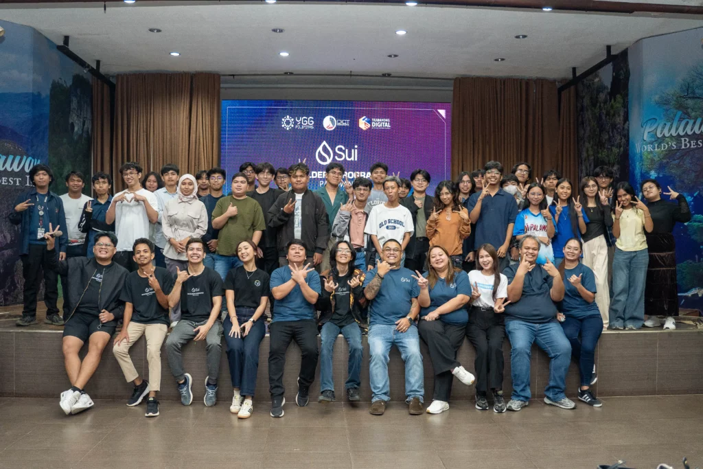 A large group of people smiling and posing for a photo on a stage. The front row is seated or crouched, consisting of a mix of men and women wearing mostly dark blue or black casual shirts. The back rows are standing. A large screen behind them displays the logos for YGG Pilipinas, DICT, and the Sui Builder Program.