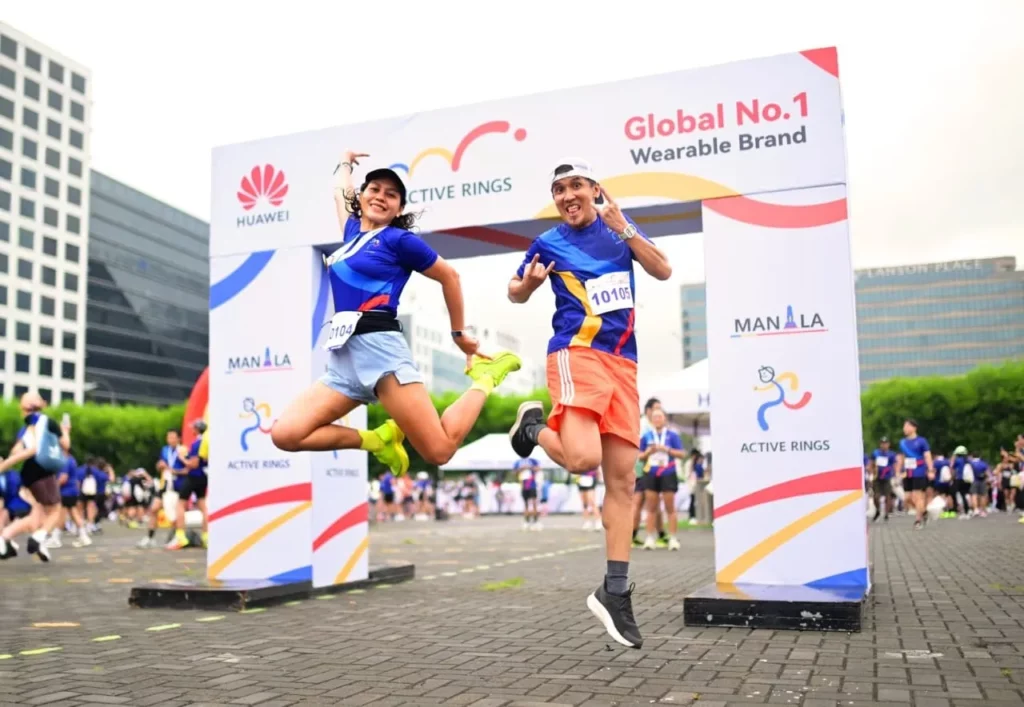 A male and a female runner jump in the air in an open outdoor area, showing excitement after the race. They are wearing race bibs 5104 and 10105, framed by a large event banner in the background displaying "Huawei Global No. 1 Wearable Brand" and "Active Rings."