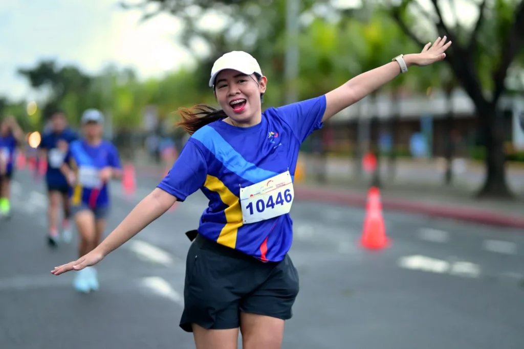 A female runner with a wide smile and a white cap is seen mid-run or celebration, facing the camera with her arms outstretched, wearing a blue and yellow shirt with race bib 10446, with the race course visible behind her.