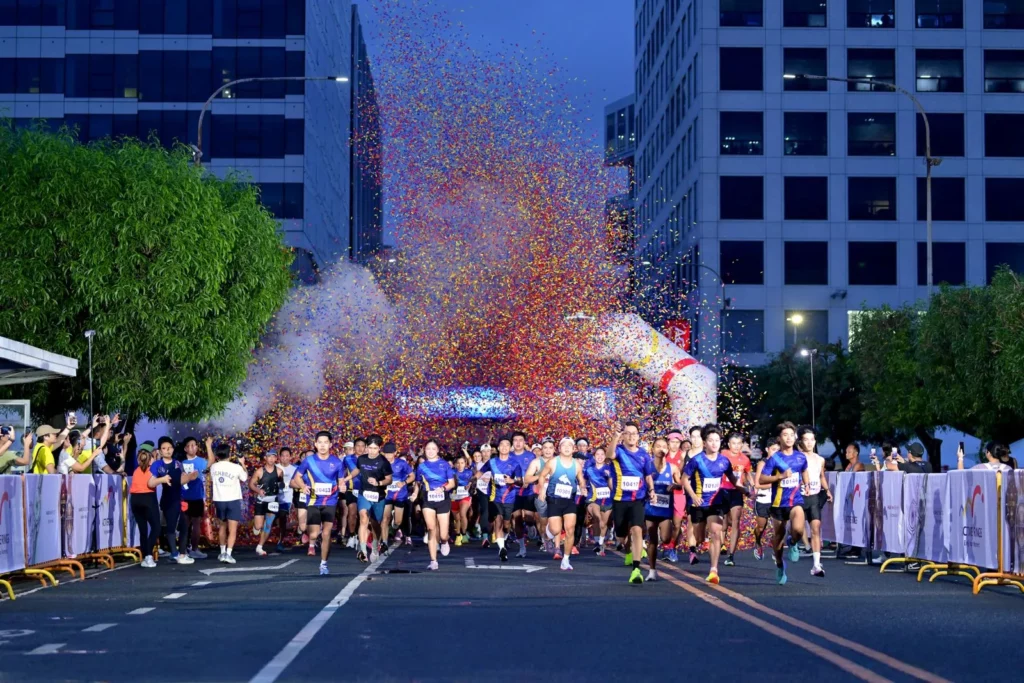 A wide shot of the start of a running race at dusk, with a large crowd of runners beginning to move forward. A huge burst of colorful confetti and smoke explodes over the inflatable start/finish archway in the background, surrounded by tall buildings.