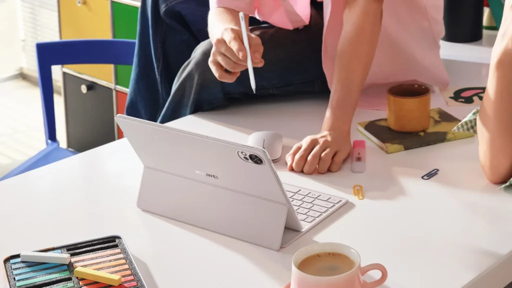 The HUAWEI MatePad 12 X 2026 set up on a white desk in its 4-in-1 workstation mode, featuring the detachable Smart Magnetic Keyboard and a wireless mouse. A hand is holding the M-Pencil stylus over the screen.