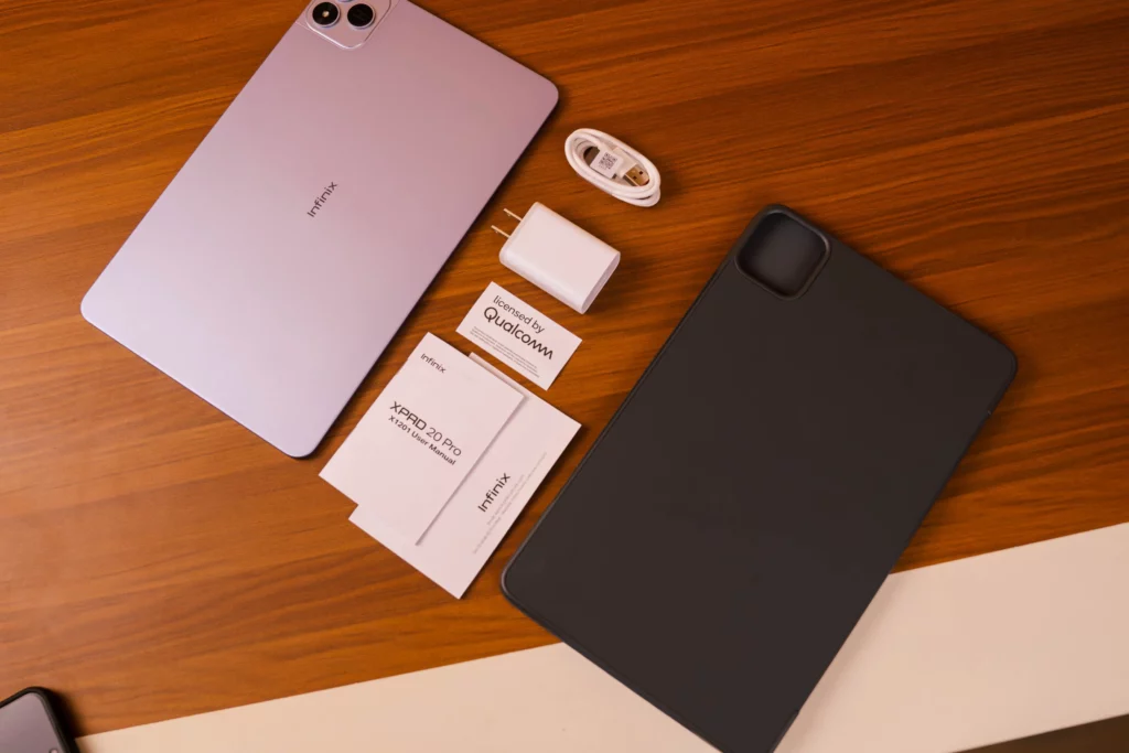 A flat lay of the Infinix XPAD 20 Pro tablet (in a lavender/light purple color) and its included accessories: a user manual, a charging adapter, a USB cable, and a protective case/cover.