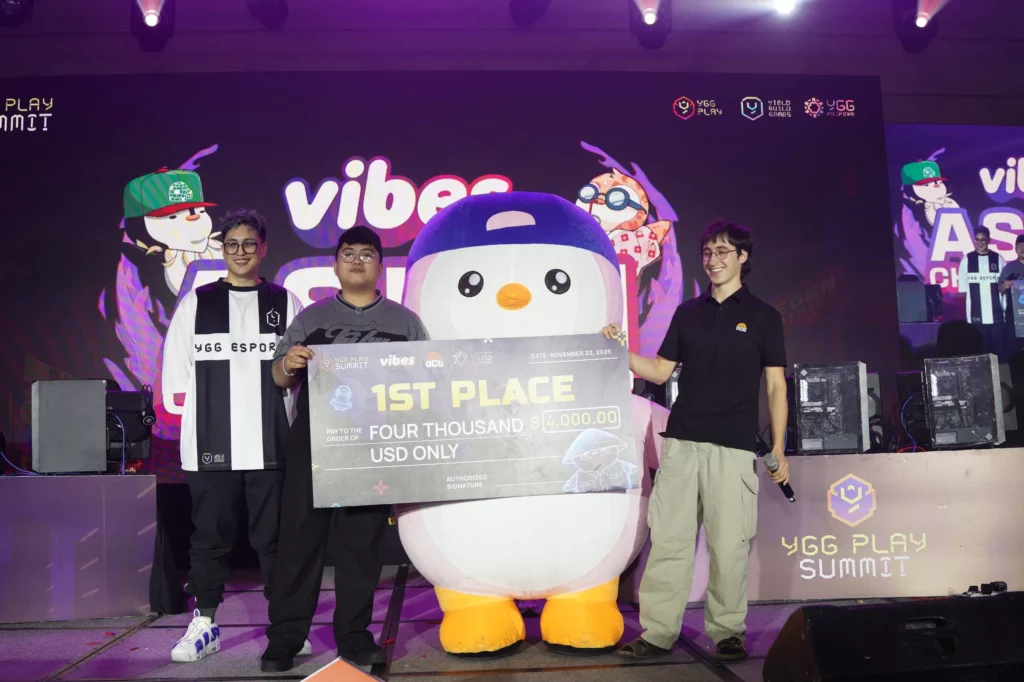 Wide shot of the Vibes Asian Championship award ceremony stage. The winner, Oxtraxex (center), holds a large check for $4,000, flanked by two other people and a mascot resembling a penguin character from the Vibes TCG. The backdrop is dark with the "YGG PLAY SUMMIT" logo visible.