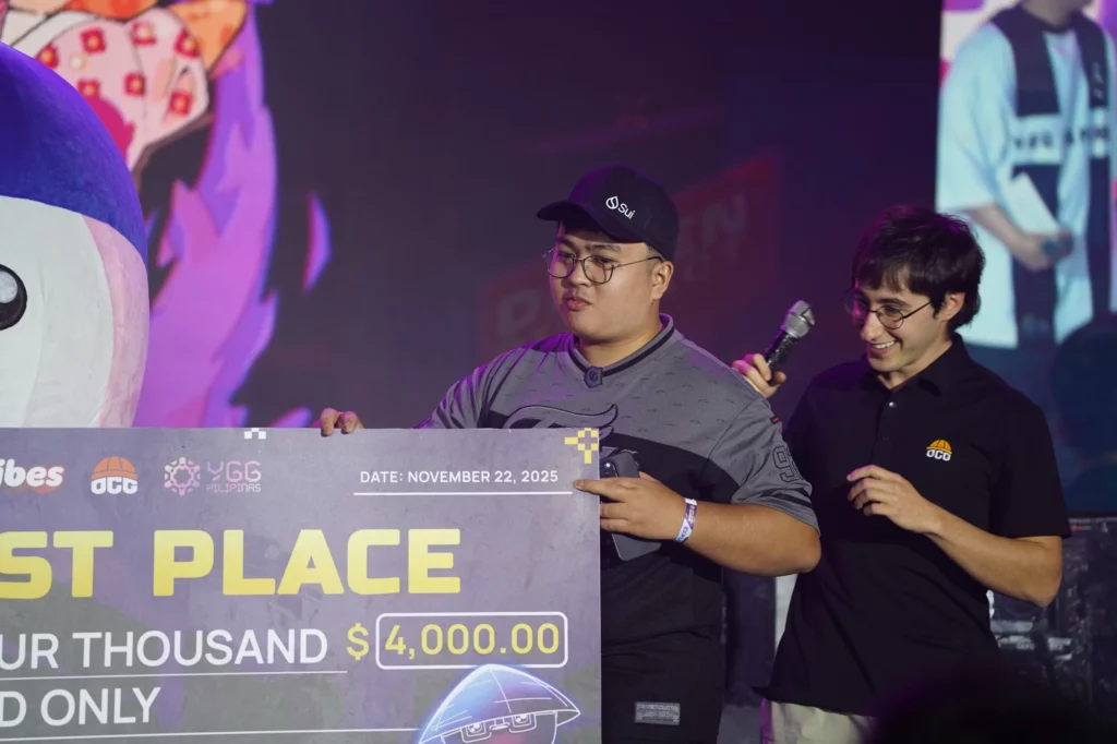 The tournament winner, a young man wearing a black cap, smiles while holding the corner of the $4,000 prize check. An event official standing beside him, holding a microphone, is also smiling. The check is dated November 22, 2025.