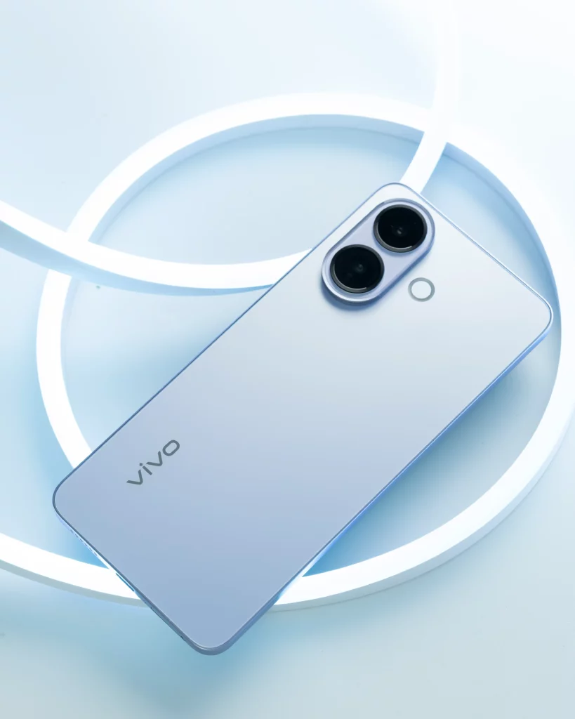 A close-up, angled shot of the rear of the blue vivo V60 Lite smartphone, highlighting the two camera lenses. The phone is set against a bright, light-blue background with glowing white neon rings.