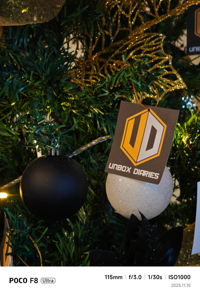 A close-up detail shot of a decorated Christmas tree. The main focus is on a matte black ornament, a sparkly white ornament, and a tag hanging from the tree that reads "UNBOX DIARIES" with a yellow and gray logo. Photo details: 115mm, f/3.0, 1/30s, ISO1000.