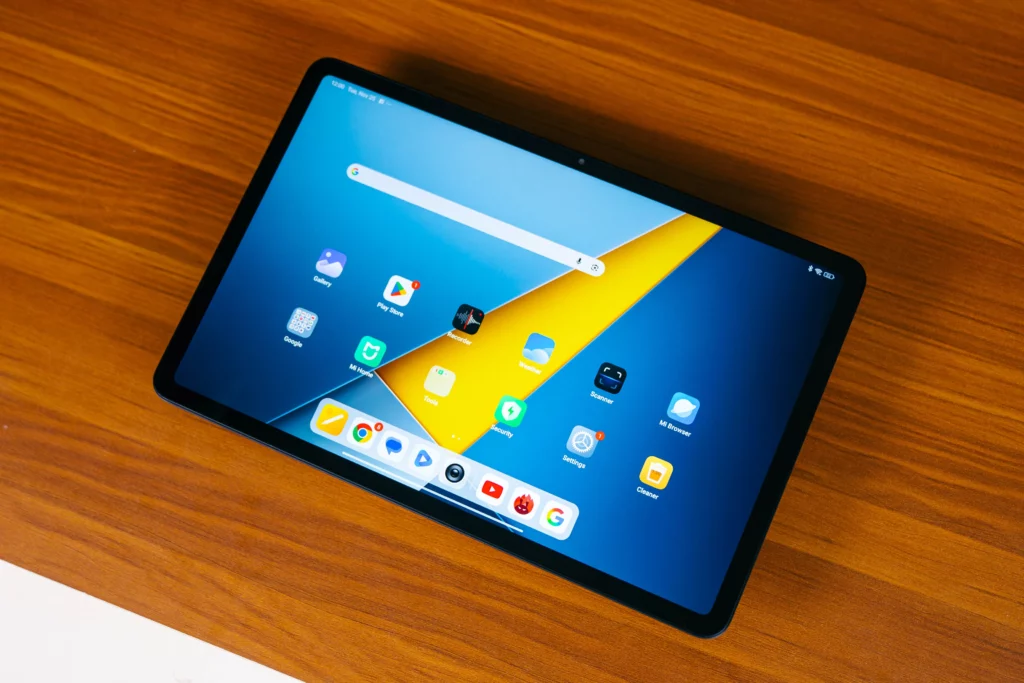 Overhead shot of the POCO Pad M1 tablet resting on a wooden table, showing its home screen with a blue and yellow wallpaper and several app icons arranged on the display.
