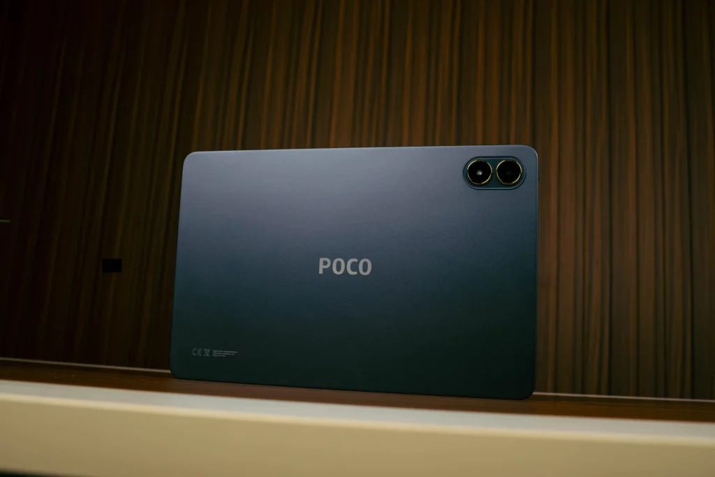 Full shot of the back of the dark gray POCO Pad M1 tablet, standing upright on a shelf. The POCO logo is centered, and the rear camera module with gold accents is visible in the top corner.