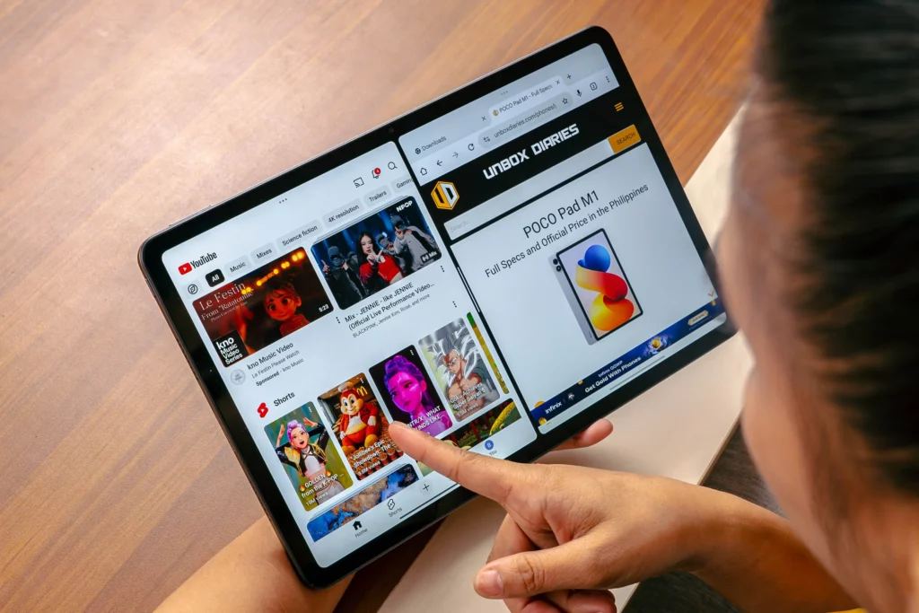An overhead shot of a person using the POCO Pad M1 tablet, demonstrating the split-screen multitasking feature with YouTube on the left and a web browser article about the POCO Pad M1 on the right. A finger is tapping the screen.