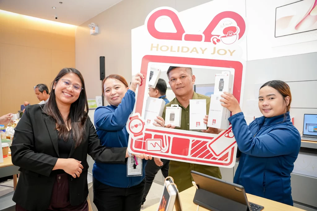 A group of four smiling people holding a large, festive "Holiday Joy" photo frame decorated with HUAWEI product icons. They are holding up boxes of HUAWEI devices, including the nova series and wearables, celebrating a purchase or event.
