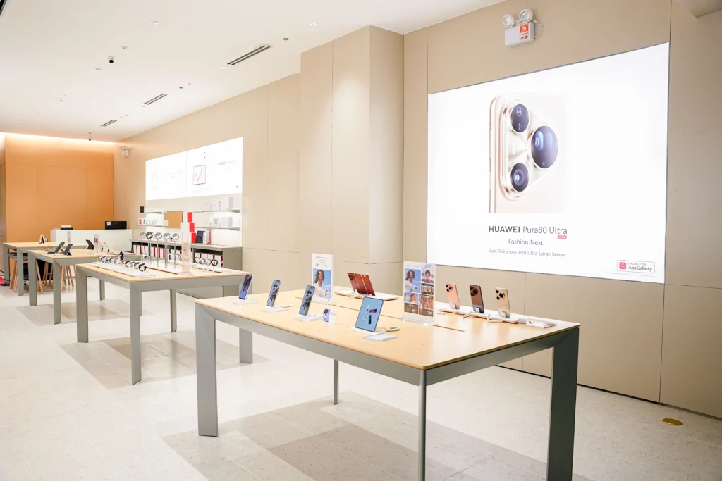 A wide-angle view of a brightly lit, modern HUAWEI Experience Store featuring minimalist wooden display tables with various smartphones and tablets. A large illuminated wall graphic showcases the HUAWEI Pura 80 Ultra with a focus on its triple-camera system.