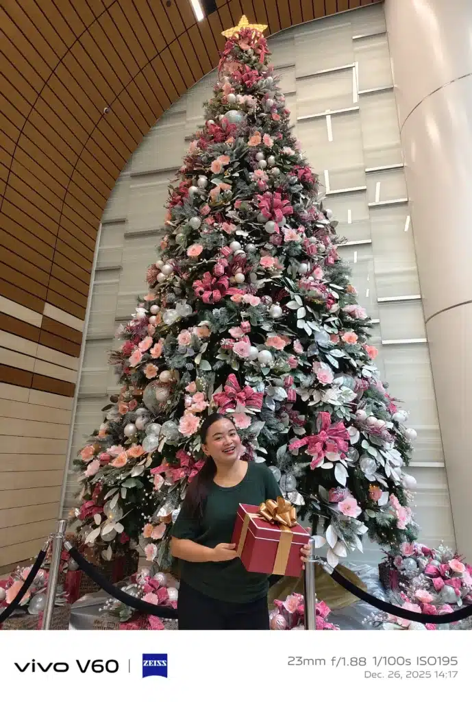 A full-length photo of a smiling woman holding a red gift box while standing in front of a towering, elaborately decorated Christmas tree in a modern indoor lobby.