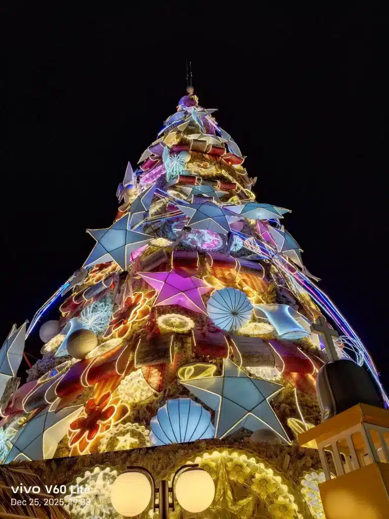 A low-angle shot of a large, brightly lit Christmas tree at night, decorated with glowing stars, shells, and colorful ornaments against a black sky.