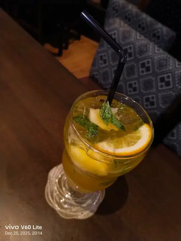 A close-up, high-angle shot of a refreshing yellow cocktail in a glass, garnished with a lemon slice and mint leaves, sitting on a wooden table.