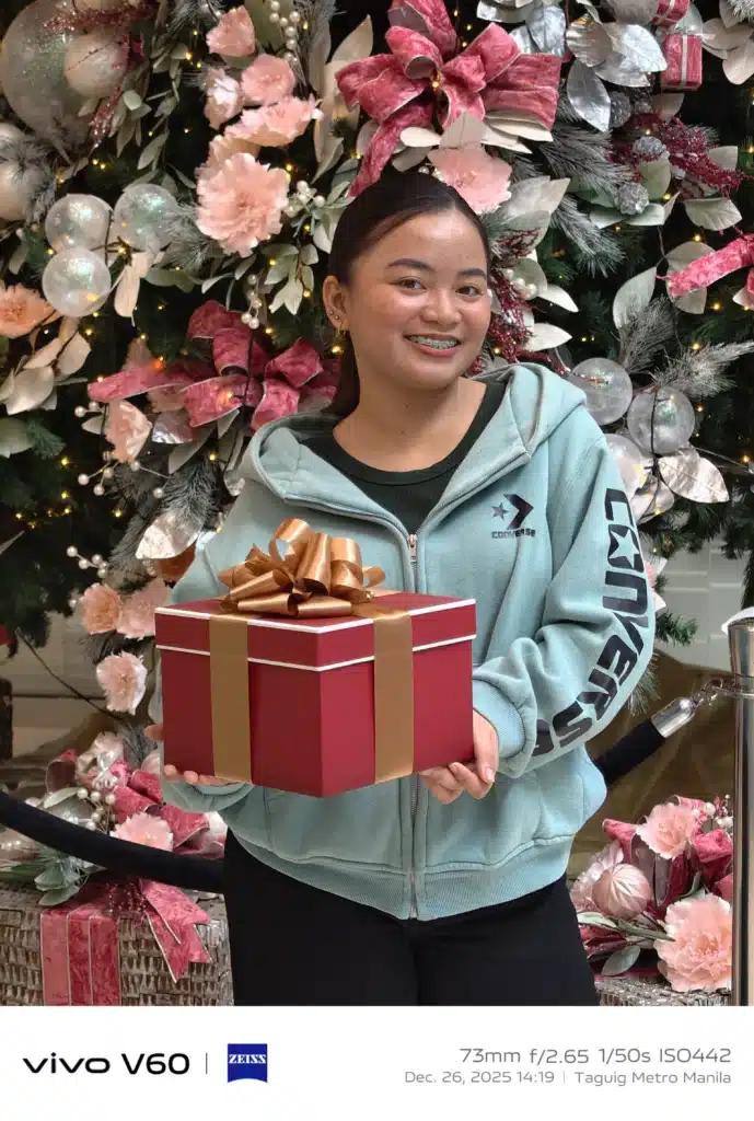 A portrait of a smiling young woman in a light blue hoodie holding a large red gift box with a gold ribbon, standing in front of a decorated Christmas tree.