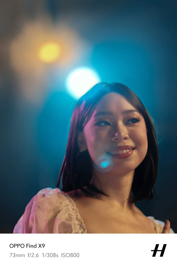 A close-up portrait of a smiling woman under atmospheric stage lighting. The image exhibits soft bokeh with glowing blue and yellow light orbs in the background, captured at f/2.6 using a 73mm lens.