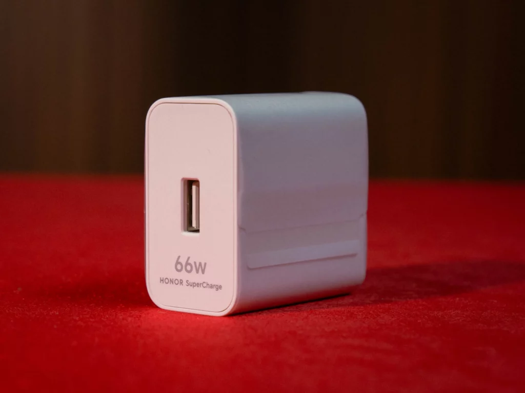 A close-up shot of a white HONOR SuperCharge power adapter sitting on a red surface. The side of the charger clearly displays "66W HONOR SuperCharge" text below a single USB-A port.