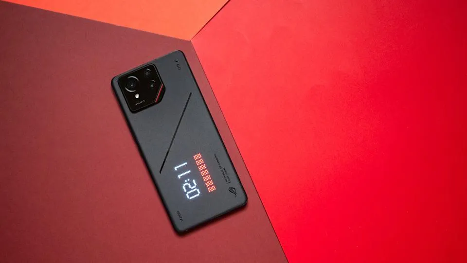 A top-down view of a black ASUS ROG Phone lying diagonally on a two-toned red and burgundy surface. The phone's rear AniMe Vision display is active, showing the digital time "02:11" in bright white LED-style pixels.