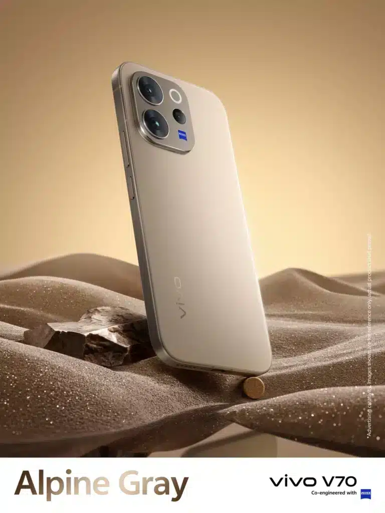 A professional product shot of the vivo V70 5G in Alpine Gray, standing upright on a textured, sand-like surface under warm lighting. The rear of the phone is visible, showcasing the triple-camera system co-engineered with ZEISS.