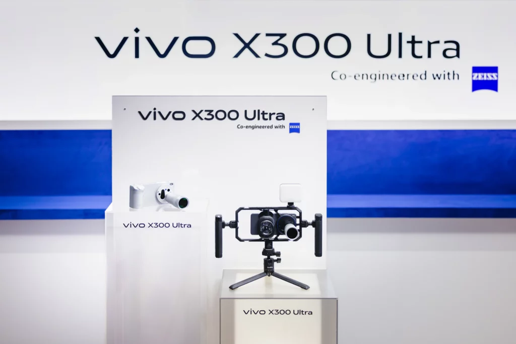 An exhibition display at MWC 2026 for the vivo X300 Ultra. A large wall sign in the background says "vivo X300 Ultra Co-engineered with ZEISS". In the foreground, two versions of the phone are displayed: one on the left with a silver telephoto lens attached, and one on the right mounted within a professional SmallRig camera cage on a tripod, complete with an external light and dual handles.