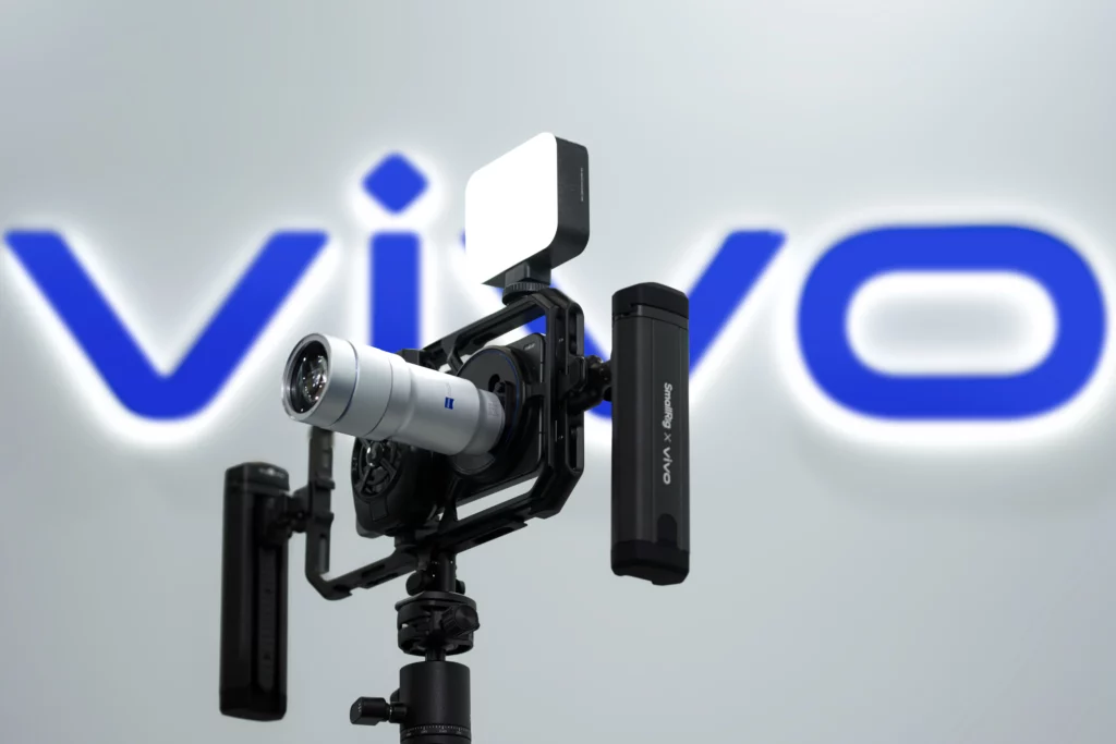A close-up shot of the professional imaging kit for the vivo X300 Ultra. The setup includes a silver ZEISS telephoto lens protruding from a black cage. The cage features side handles labeled "SmallRig x vivo" and a square LED light mounted on top. The entire rig is mounted on a tripod against a blurred background with a glowing vivo logo.