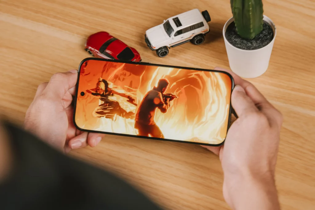 The POCO X8 Pro Iron Man Edition being held by two hands with two model cars and a succulent at the back. The POCO X8 Pro's screen showing some kind of animated graphic.