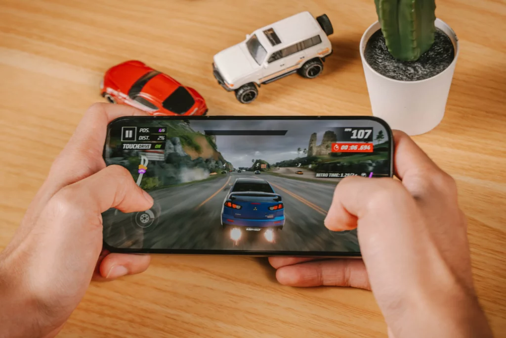 The POCO X8 Pro Iron Man Edition being held by two hands with two model cars and a succulent at the back. The photo showing someone playing a fast-action driving game in the POCO X8 Pro.