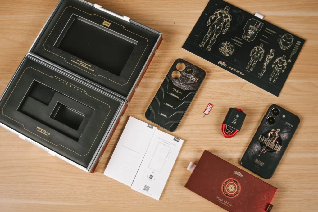 The unboxed contents of the POCO X8 Pro Iron Man Edition spread out on a table. Includes the smartphone, a custom black protective case with Tony Stark's signature, a red USB-C charging cable, and a themed SIM ejector tool shaped like a Marvel logo.