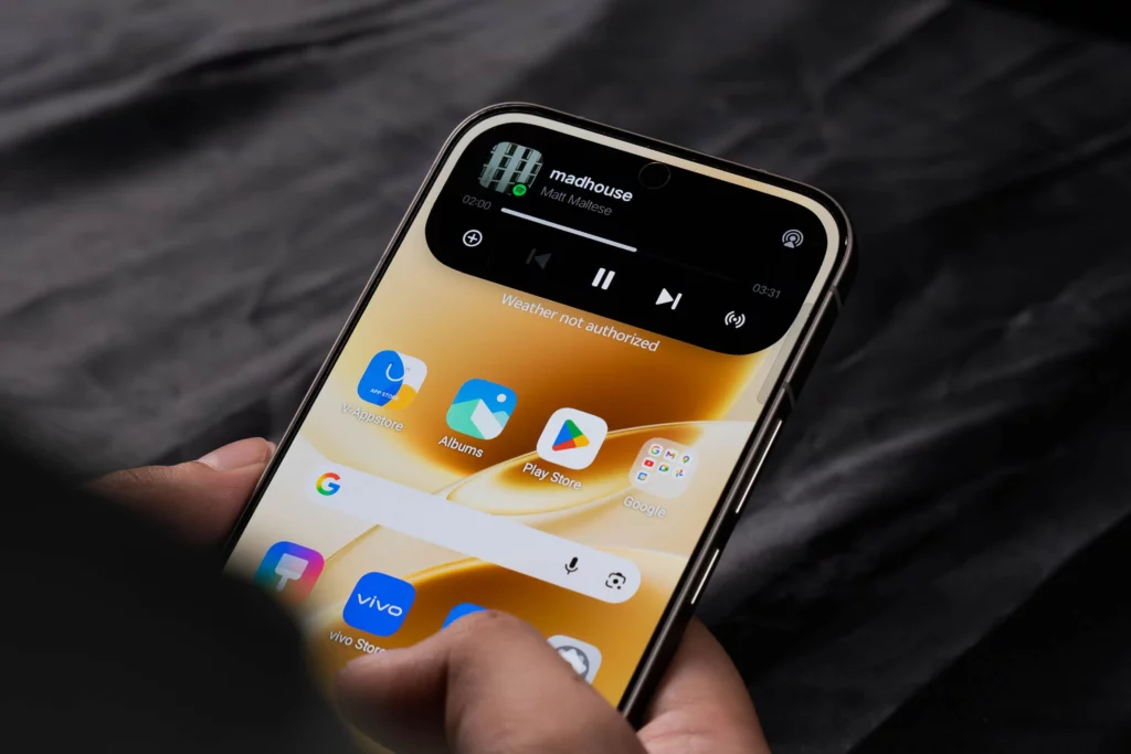 A person holding the vivo V70 5G showing the home screen. A large "Dynamic Island" style music widget is active at the top, displaying the song "Madhouse" by Matt Maltese.