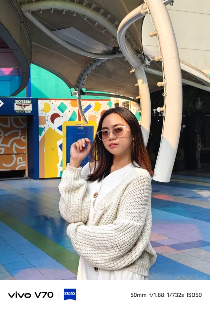 A portrait photo of a woman wearing glasses and a white cardigan, taken with the vivo V70 5G. The bottom watermark indicates it was shot using the ZEISS co-engineered camera at 50mm, f/1.88.