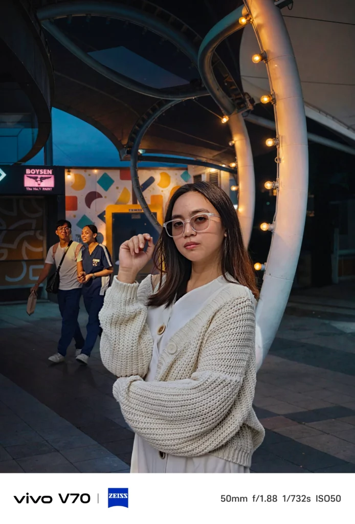 A portrait shot of the same woman at dusk, showing the camera's ability to capture warm ambient light from a nearby circular light fixture.