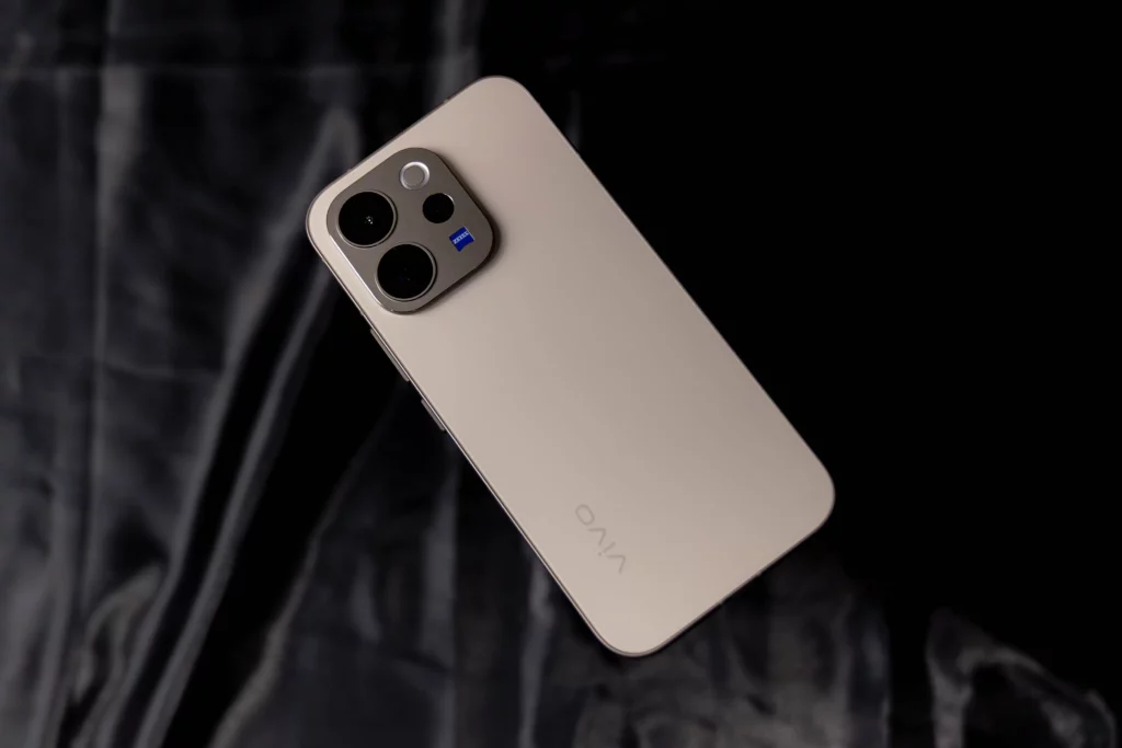 A top-down view of the vivo V70 5G resting on a dark, textured silk-like fabric. The elegant cream-colored back and the prominent ZEISS-branded camera module are clearly visible.