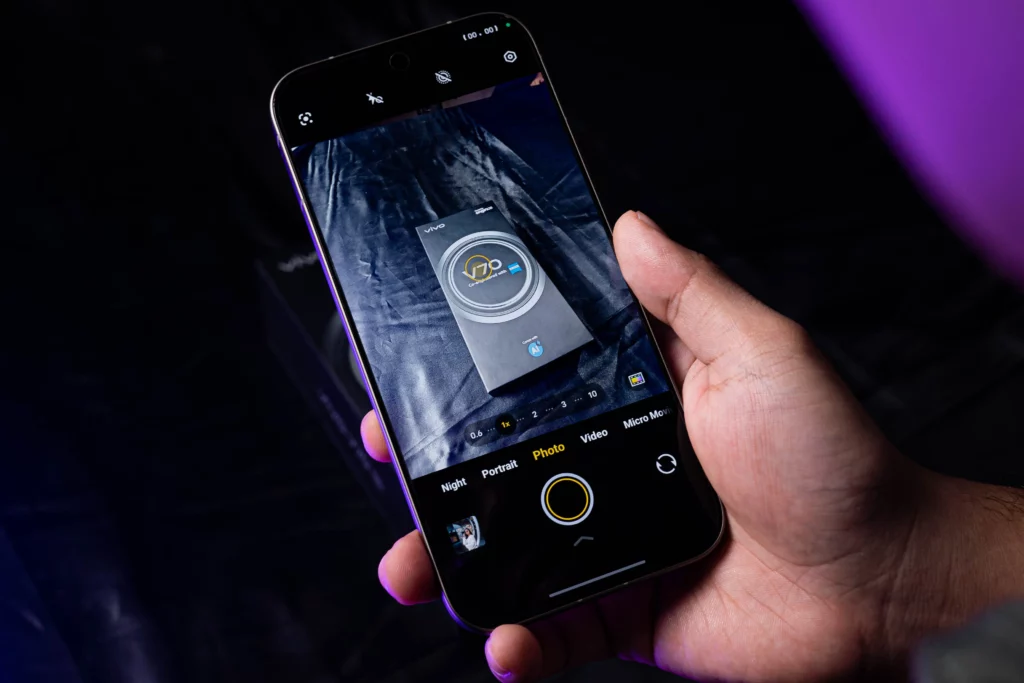 A person holding the vivo V70 5G with the screen on, showing the camera app interface. The viewfinder is capturing the phone's own retail box, showcasing the thin bezels and center punch-hole selfie camera.