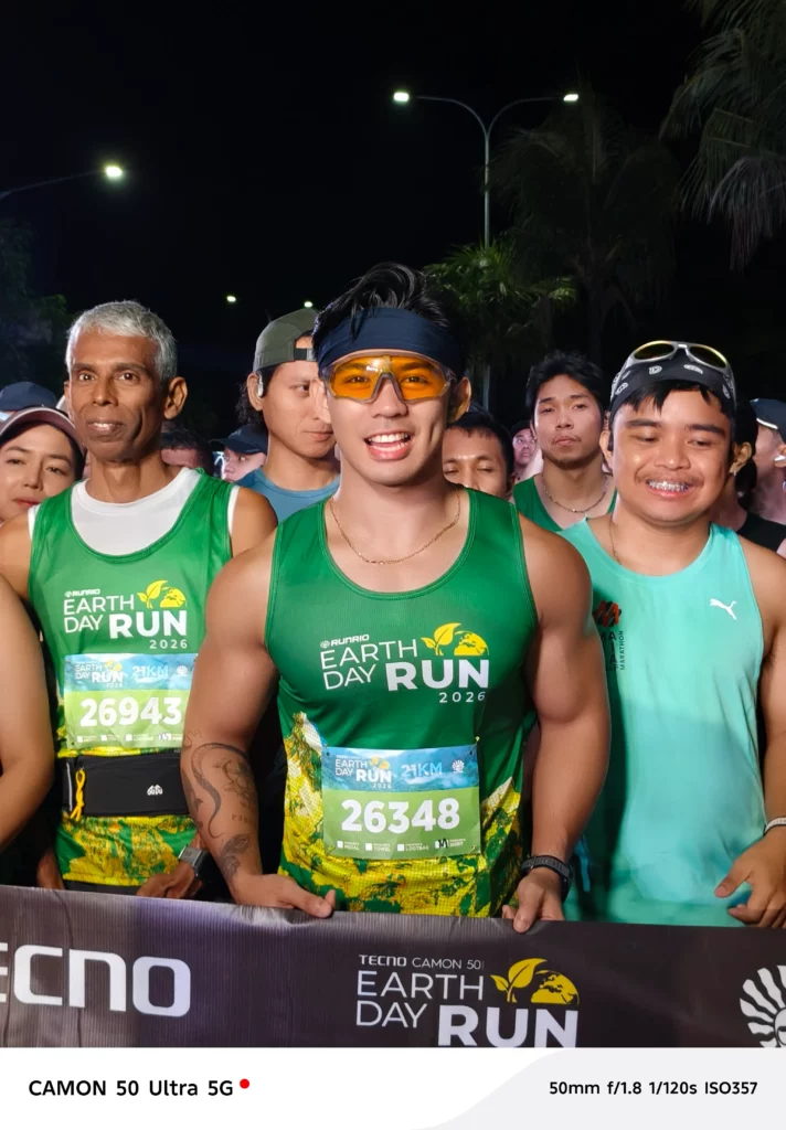 A close-up portrait of a male runner in a green Earth Day Run singlet and yellow-tinted sports glasses, smiling brightly while surrounded by other participants at the starting line.
